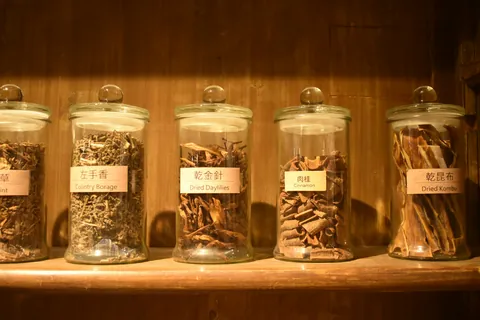 Glass jars of dried herbs used in traditional Chinese medicine on a wooden shelf